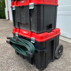 May include: A stack of black and red toolboxes with silver latches. A green extension cord is attached to the side. The bottom toolbox has a wheel for easy transport. The toolboxes are stacked on a concrete surface.