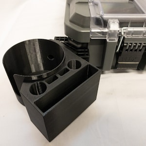 Peut inclure: Un organisateur noir imprimé en 3D avec un porte-gobelet, deux petits supports circulaires et un compartiment rectangulaire. L'organisateur est placé à côté d'un conteneur de stockage gris avec un couvercle transparent et un loquet noir. Le logo FLEX est visible.