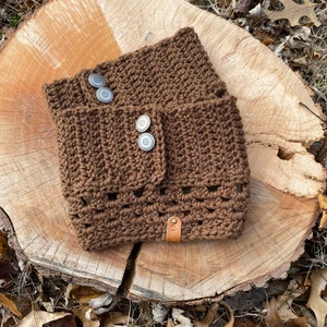 May include: A brown crocheted neck warmer with a leather tag and silver buttons. The warmer is sitting on a wooden surface.