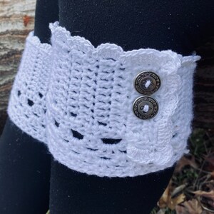 May include: White crocheted leg warmer with a scalloped edge and two silver buttons. The leg warmer is worn over black leggings.