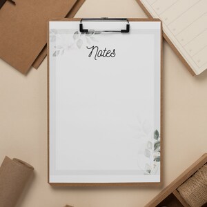 May include: A clipboard with a white sheet of paper that says "Notes" at the top. The paper has a watercolour design of leaves and flowers. The clipboard is on a beige surface with other stationery.