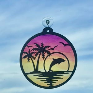 May include: A round, black silhouette of a tropical beach scene with a dolphin leaping from the water. The background features a sunset with pink, orange, and yellow hues. Palm trees and birds are also silhouetted.
