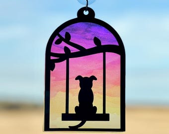 May include: A black silhouette of a dog sitting on a swing against a sunset background. The swing is hanging from a branch with leaves. The background is a gradient of purple, pink, orange, and yellow. The image is framed in a black border and has a suction cup at the top.