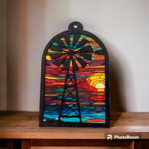 May include: A wooden cutout of a windmill with a stained glass design. The windmill is silhouetted against a sunset with orange, red, and blue hues. The background is a light gray wall.