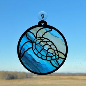 Op de afbeelding: Een rond, zwart ingelijst glas-in-loodraam dat hangt met een zuignap. Het glas-in-lood toont een blauwe en groene zeeschildpad die zwemt in een blauwe en witte abstracte achtergrond.