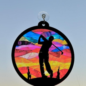 May include: A round, black, silhouette of a golfer swinging a club against a colorful, stained glass background. The background features a sunset with shades of blue, pink, orange, and yellow.