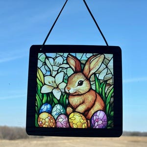 以下が含まれることがあります： カラフルなイースターエッグと白い花に囲まれた茶色のウサギが描かれたステンドグラスの窓が吊り下げられています