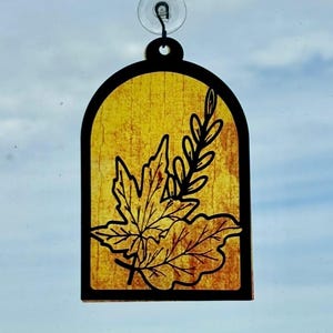 May include: A decorative window cling featuring a leaf design. The cling has a black border and a yellow background. The leaf design is black and includes a maple leaf and a sprig of leaves. A suction cup is attached to the top.