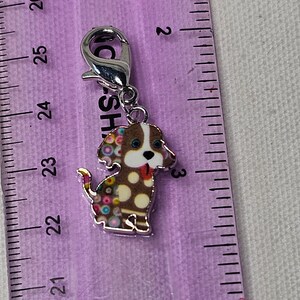Pu&ograve; includere: Un ciondolo d'argento con un cane marrone e bianco con pois colorati. Il cane ha la lingua rosa che sporge e gli occhi blu. Il ciondolo ha una chiusura argentata.
