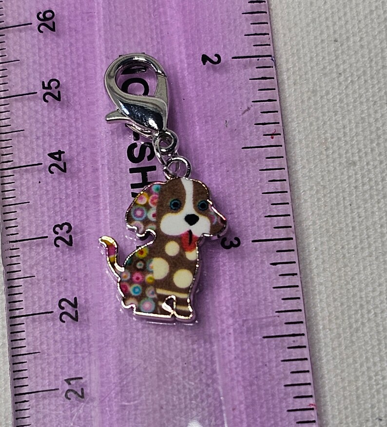 Pu&ograve; includere: Un ciondolo d'argento con un cane marrone e bianco con pois colorati. Il cane ha la lingua rosa che sporge e gli occhi blu. Il ciondolo ha una chiusura argentata.