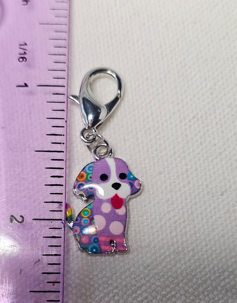 Pu&ograve; includere: Un ciondolo a forma di cane viola e bianco con pois colorati e una chiusura argentata. Il ciondolo &egrave; alto circa 2,5 cm.