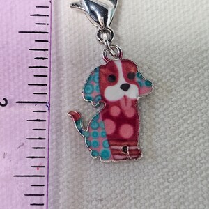 Pu&ograve; includere: Un ciondolo d'argento con un design di cane rosso, bianco e blu. Il cane ha una faccia bianca con occhi neri e un naso nero. Il cane indossa una camicia con un motivo blu e rosa.