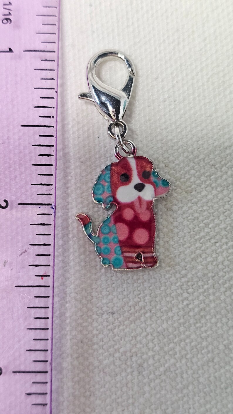 Pu&ograve; includere: Un ciondolo d'argento con un design di cane rosso, bianco e blu. Il cane ha una faccia bianca con occhi neri e un naso nero. Il cane indossa una camicia con un motivo blu e rosa.