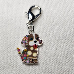 Pu&ograve; includere: Un ciondolo d'argento con un cane marrone e bianco con pois colorati. Il cane ha gli occhi azzurri e la lingua rosa che sporge. Il ciondolo ha una chiusura a moschettone argentata.