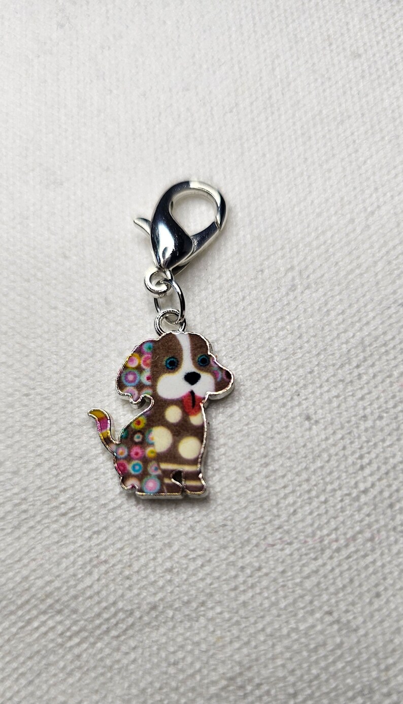 Pu&ograve; includere: Un ciondolo d'argento con un cane marrone e bianco con pois colorati. Il cane ha gli occhi azzurri e la lingua rosa che sporge. Il ciondolo ha una chiusura a moschettone argentata.