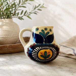 May include: A cream-colored ceramic pitcher featuring a hand-painted design with deep blue, yellow, and green floral motifs. The body displays a central yellow oval surrounded by a dark blue sunburst pattern, with additional blue leaf accents.