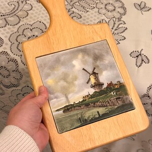 May include: Wooden chopping board with a handle and a rectangular tile insert. The tile depicts a landscape painting of a windmill, water, and buildings. The wood is light-coloured.