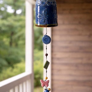 May include: A blue ceramic wind chime with a bell-shaped top featuring floral and circular patterns. The chime has a series of decorative elements, including a pink butterfly, a blue sphere, and various beads. The background shows a porch and greenery.