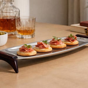 May include: A silver serving platter with dark wooden legs displays four appetizers. Each appetizer is a cracker topped with meat and a sprig of herb. Whiskey and olives are in the background, creating a sophisticated setting.