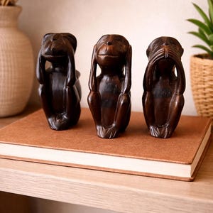 May include: Three dark brown wooden monkey figurines, each in a different pose representing "See no evil, hear no evil, speak no evil." The figurines are displayed on a brown book on a wooden shelf. The background includes a vase with dried flowers and a potted plant.