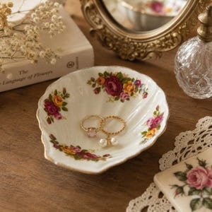 May include: A white ceramic shell-shaped trinket dish featuring a delicate floral pattern of red, yellow, and pink roses with green leaves. The scalloped edges are trimmed in gold. Two gold-toned rings rest inside the dish, one with a pink stone.