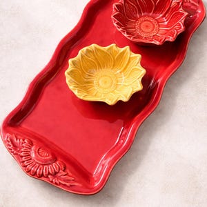 May include: A red ceramic serving tray with a wavy edge, featuring a sunflower design. Two sunflower-shaped bowls, one red and one yellow, sit on the tray. The tray has a sunflower design on the end.