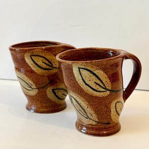 Puede incluir: Dos tazas de cerámica con un esmalte marrón moteado y un borde acampanado. Cada taza presenta un patrón pintado a mano de hojas de color crema delineadas en marrón oscuro. Las tazas tienen un asa curva y una base de pedestal.