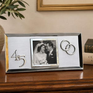 May include: A rectangular silver-toned metal picture frame featuring a brushed finish, a central square photo of a couple, the silver number 45 on the left, and two interlocking silver rings on the right side of the frame's front surface.