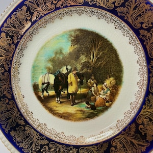 May include: A decorative ceramic plate with a detailed pastoral scene in the center. The scene depicts figures with horses and a dog. The plate has a white background with a blue and gold ornate border.