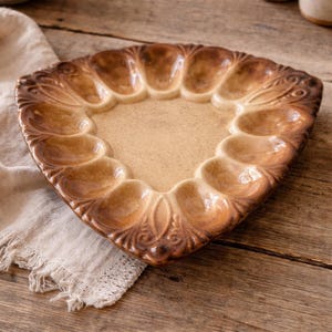 Puede incluir: Una fuente de cerámica marrón para servir huevos con un borde decorativo festoneado. La fuente tiene 12 hendiduras en forma de huevo y un área central. El color es un marrón cálido y terroso con tonos beige claro en el centro. La fuente está sobre una superficie de madera.