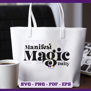 Könnte beinhalten: Weiße Einkaufstasche mit schwarzem Text, der "Manifest Magic Daily" lautet. Die Tasche liegt auf einem Tisch mit einer Kaffeetasse und einem Stapel Büchern.