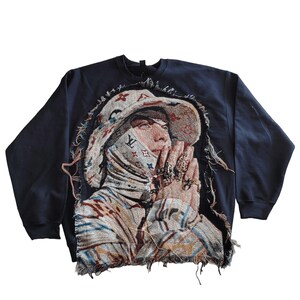 May include: A navy blue sweatshirt with a tapestry design featuring a person wearing a hat and face covering. The tapestry has a beige, brown, and blue color scheme. The edges of the sweatshirt are frayed.