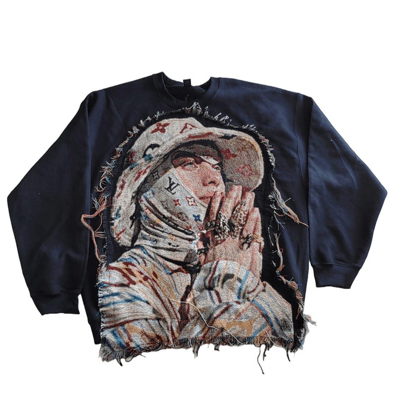 May include: A navy blue sweatshirt with a tapestry design featuring a person wearing a hat and face covering. The tapestry has a beige, brown, and blue color scheme. The edges of the sweatshirt are frayed.