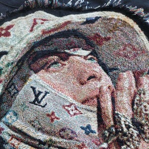 May include: Woven tapestry blanket featuring a portrait of a person with their hands near their face. The blanket has a multi-colored design with the letter "W" and other symbols. The edges of the blanket are fringed.