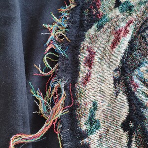 May include: Close-up of a black textile with a tapestry design. The tapestry features a mix of colors, including green, red, and black. The edge of the textile has colorful, frayed threads.