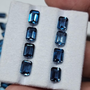 May include: Eight rectangular, deep blue gemstones are arranged on a white surface. The gems have a polished, reflective surface, and are likely cut for jewelry. The gems are a rich, saturated blue color.