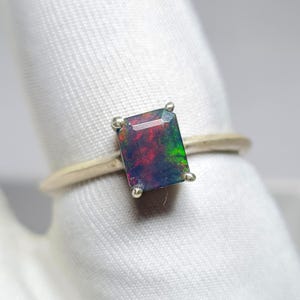 Puede incluir: Anillo de plata con una piedra preciosa de ópalo negro rectangular que muestra colores rojos y verdes. El ópalo está sujeto por cuatro pequeñas garras, y la banda es de plata pulida.