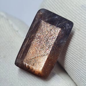 May include: A rectangular gemstone with a polished surface. The gem has brown, gold, and iridescent colours. The stone's facets catch the light, creating a sparkling effect. The gemstone is likely used for jewellery.