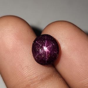 以下が含まれることがあります： 深い紫色のスターサファイアの宝石。 宝石は丸みを帯びた形状で、表面に星のような効果が見られます。 色は、微妙な光沢のある、豊かで濃い紫色です。 宝石は2本の指の間に挟まれています。