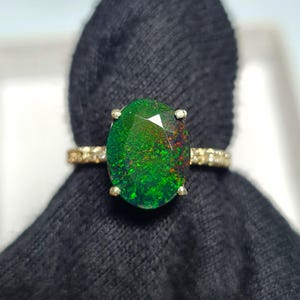 May include: A gold ring featuring a large, oval-cut opal with vibrant green and red internal colors. The ring's band is adorned with small, sparkling accents. The opal is set with four prongs, showcasing its unique color play.