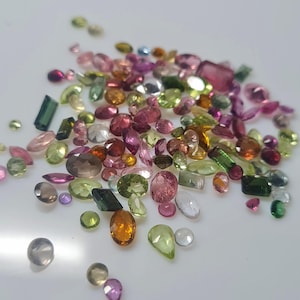 Può includere: Una collezione di gemme colorate di varie forme e dimensioni. Le gemme includono sfumature di rosa, verde, arancione e trasparente, con tagli ovali, a goccia e rettangolari. Le gemme sono sparse su una superficie bianca.