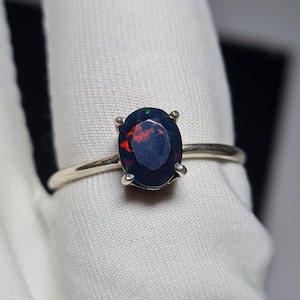 Puede incluir: Un anillo de plata con una piedra preciosa de ópalo ovalada oscura. El ópalo muestra destellos rojos y verdes dentro de su cuerpo azul-negro oscuro. La banda del anillo es un diseño simple y delgado de plata, que resalta la gema.
