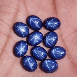 Könnte beinhalten: Nahaufnahme von neun ovalen, tiefblauen Edelsteinen. Jeder Stein zeigt ein markantes sechsstrahliges Sternmuster in einem helleren Blau, das einen auffälligen Kontrast erzeugt. Die Steine sind glatt und poliert und reflektieren das Licht.