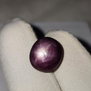 Puede incluir: Una gema ovalada pulida, de color púrpura intenso. La piedra exhibe un efecto de estrella, con líneas que irradian desde un punto central. La gema se sostiene sobre un fondo de tela blanca, resaltando su color y brillo.