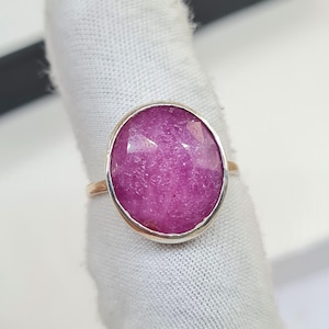 Puede incluir: Un anillo de plata con una gran gema rosa ovalada y facetada. La gema está engastada en un bisel de plata. El anillo se muestra sobre una tela blanca. La superficie de la gema es brillante.