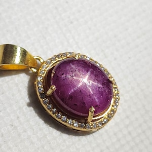 May include: A gold pendant featuring a large, oval-shaped, purplish-red gemstone with a star-like pattern. The gemstone is surrounded by a halo of small, clear stones and set in a gold frame. The pendant has a gold bail.