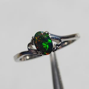 Könnte beinhalten: Ein silberner Ring mit einem ovalen, schwarzen Opal-Edelstein, der grüne und rote Lichtreflexe zeigt. Der Opal ist in einem zarten, wirbelnden Silberband gefasst. Das Design des Rings ist elegant und auffällig. Der Hintergrund ist weiß.