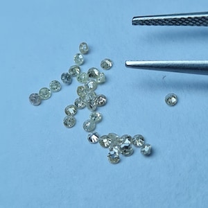May include: A collection of small, round, faceted diamonds scattered on a light blue surface. The diamonds vary in color, with some appearing clear and others with a slight yellow tint. A pair of tweezers is positioned above the diamonds.