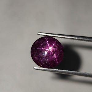 Op de afbeelding: Een ronde, gepolijste, dieppaarse edelsteen, vastgehouden door een pincet. De edelsteen heeft een zespuntig sterpatroon dat vanuit het midden uitstraalt. Het stereffect wordt gecreëerd door lichtreflectie van insluitsels in de steen. De edelsteen is een cabochon-slijpsel.