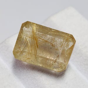 Pode incluir: Uma gema retangular e transparente com inclusões douradas em forma de agulha. A gema tem uma superfície polida e uma cor amarelo-castanho claro. A gema está sobre um fundo branco.